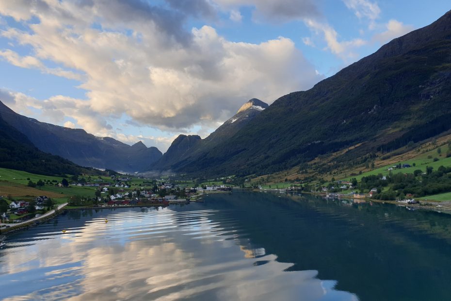 A blue sky with tall clouds is reflected on the still waters. Large hills and mountains frame the water, and near the shore, green fields and clusters of buildings.