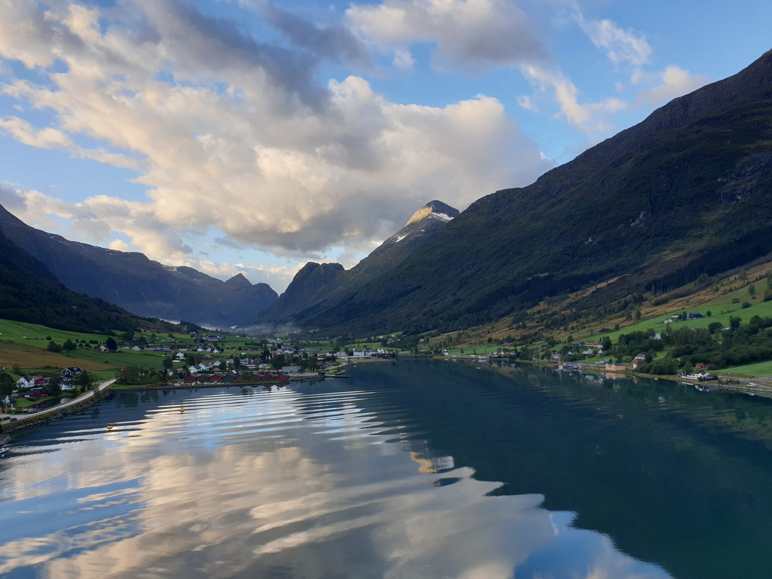 A blue sky with tall clouds is reflected on the still waters. Large hills and mountains frame the water, and near the shore, green fields and clusters of buildings.