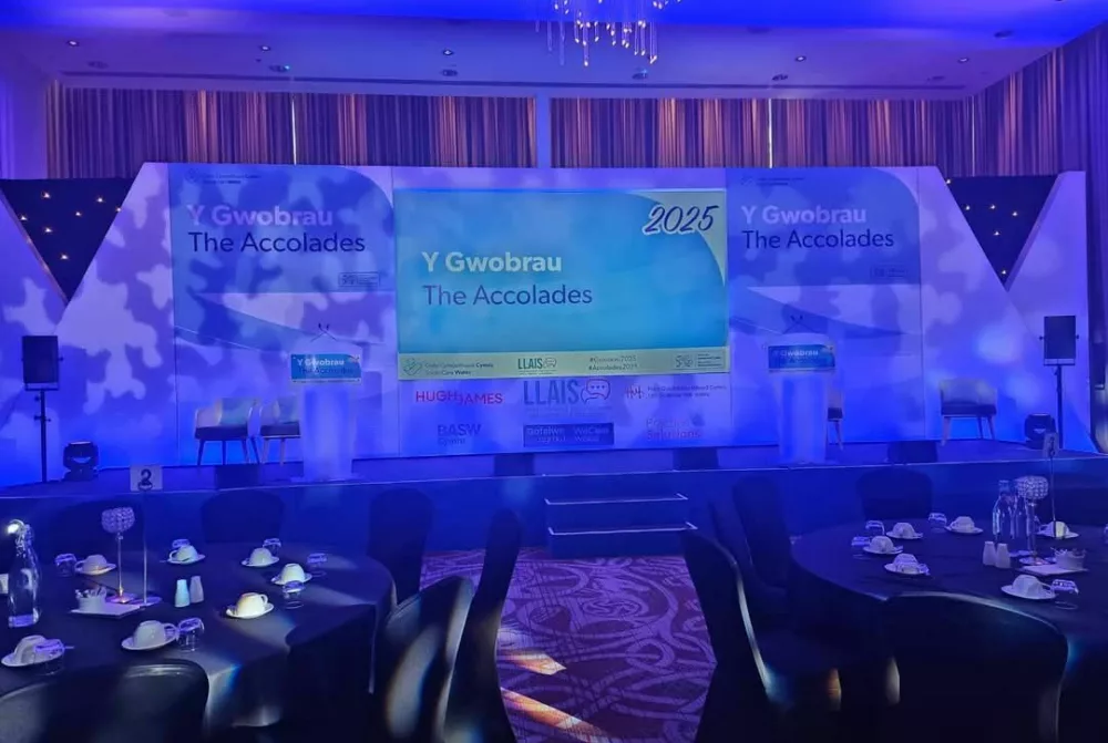 The Accolades stage setup before the room filled with people! There are round tables with black tablecloths in the foreground, and the stage has a display screen surrounded by sponsor logos and the Accolades logo. Everything is bathed in purple lighting.