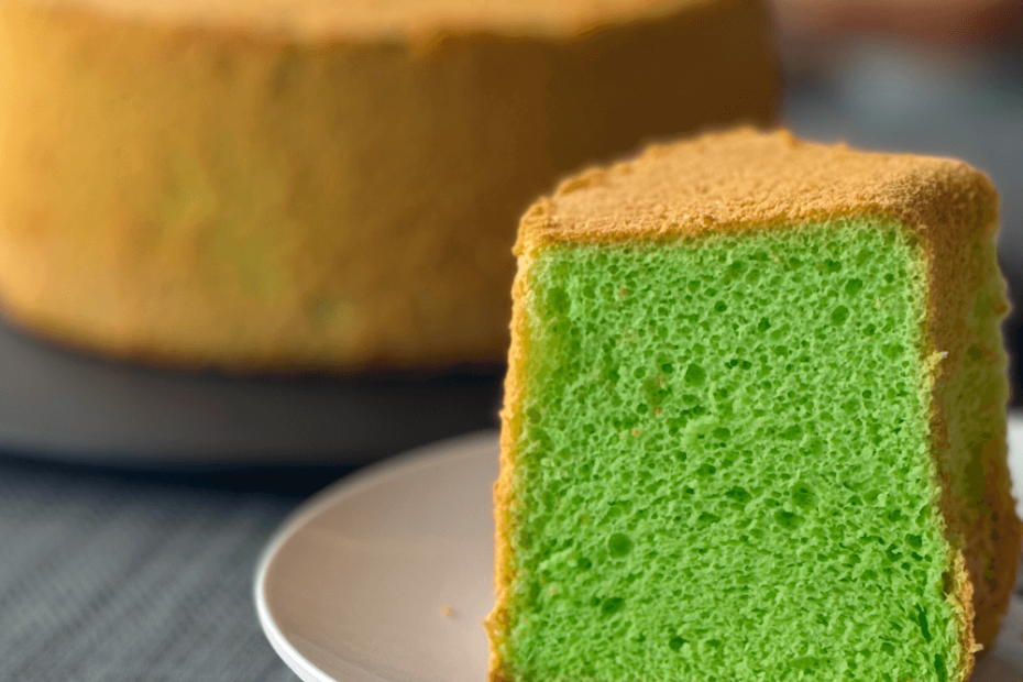 A fresh green hued thick slice of pandan chiffon cake on a plate; in the background the rest of the cake on a display stand.