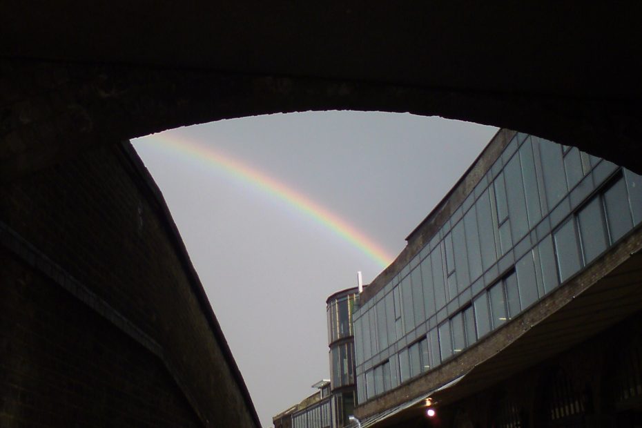 A curved triangular shape is created by an archway and a wall in front of a glass building. Everything is in tones of dark greys and black, but the triangle of light sky is bisected by a thin but sharp rainbow.