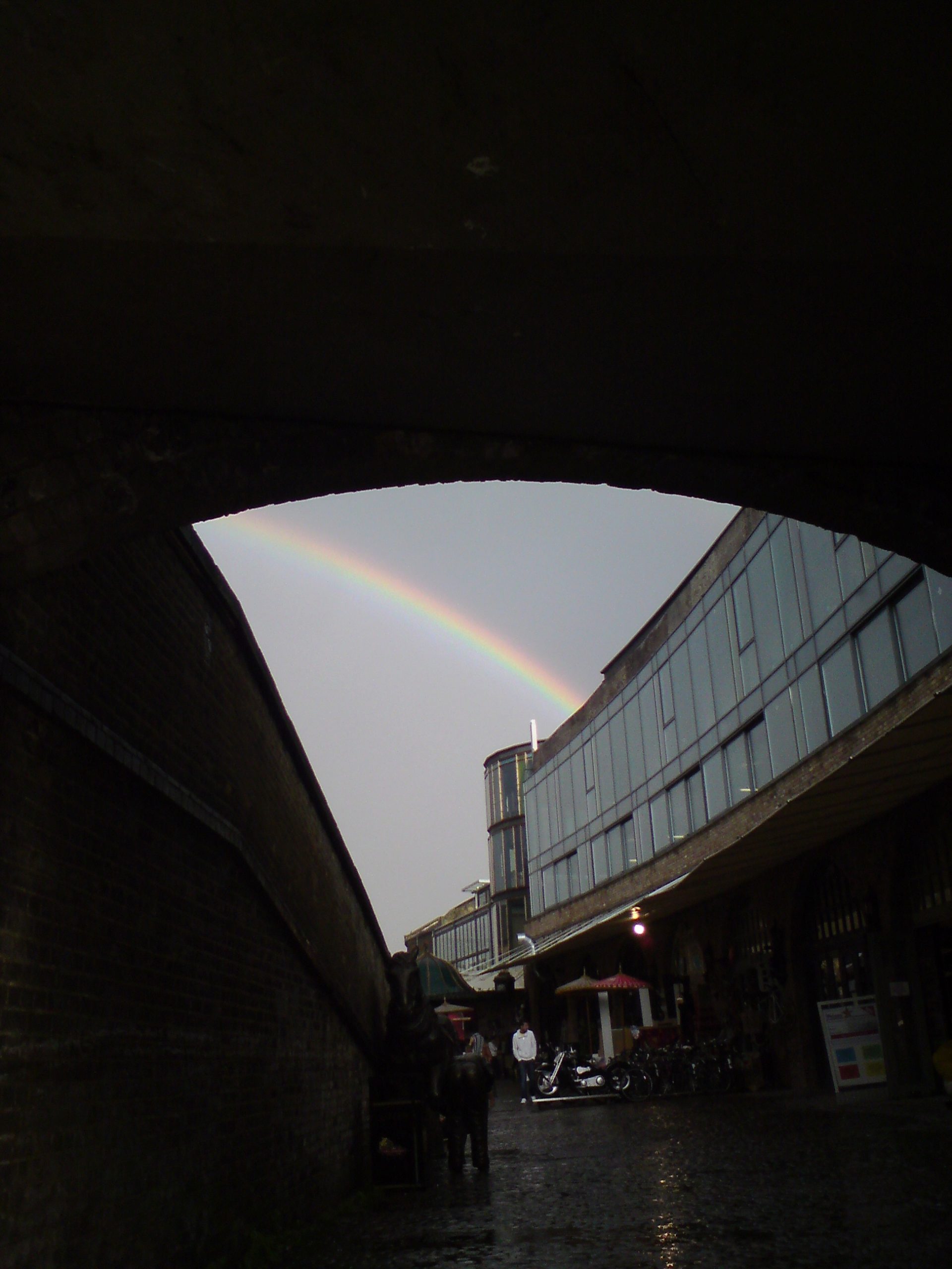 A curved triangular shape is created by an archway and a wall in front of a glass building. Everything is in tones of dark greys and black, but the triangle of light sky is bisected by a thin but sharp rainbow.