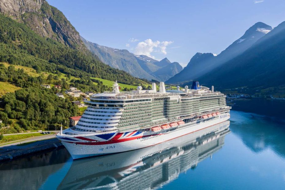 A massive cruise ship is docked at the edge of a tranquil fjord, hills and mountains all around.
