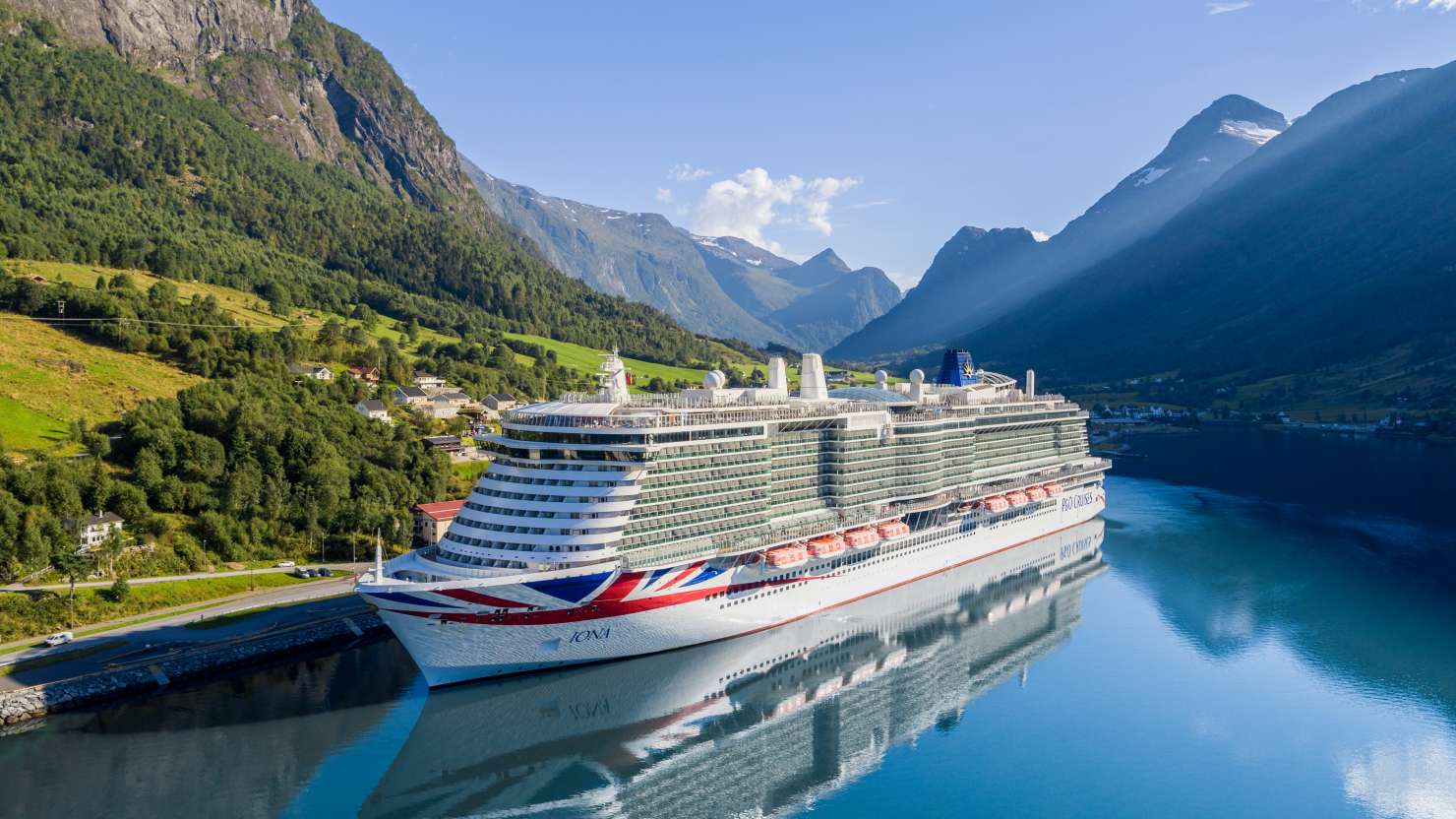 A massive cruise ship is docked at the edge of a tranquil fjord, hills and mountains all around.