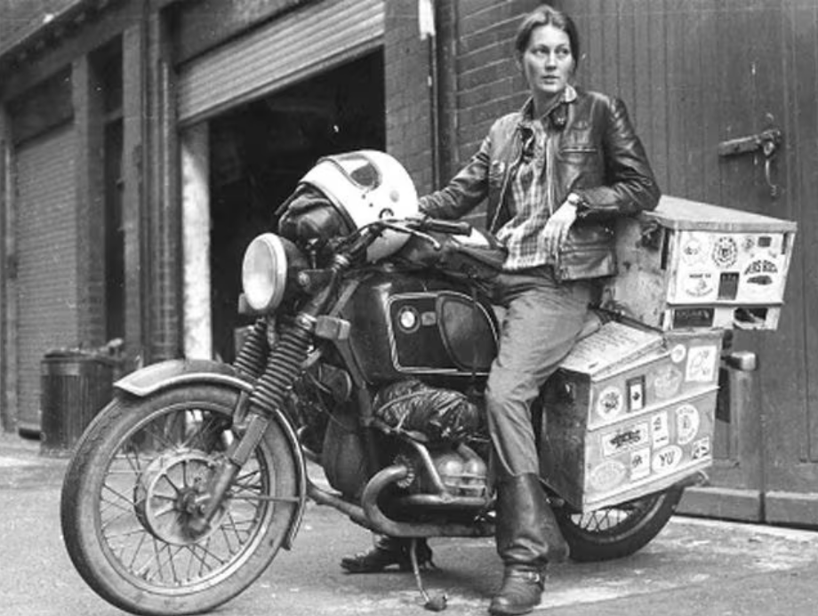 Blach and white photo. Elspeth Beard astride her BMW motorbike, with large side and rear panniers (designed and built by her out of sheet aluminium and rivets), with a white helmet resting on the handlebars. She's wearing jeans, leather boots, and a leather jacket. She's parked up outside what looks like a warehouse, looking off camera.