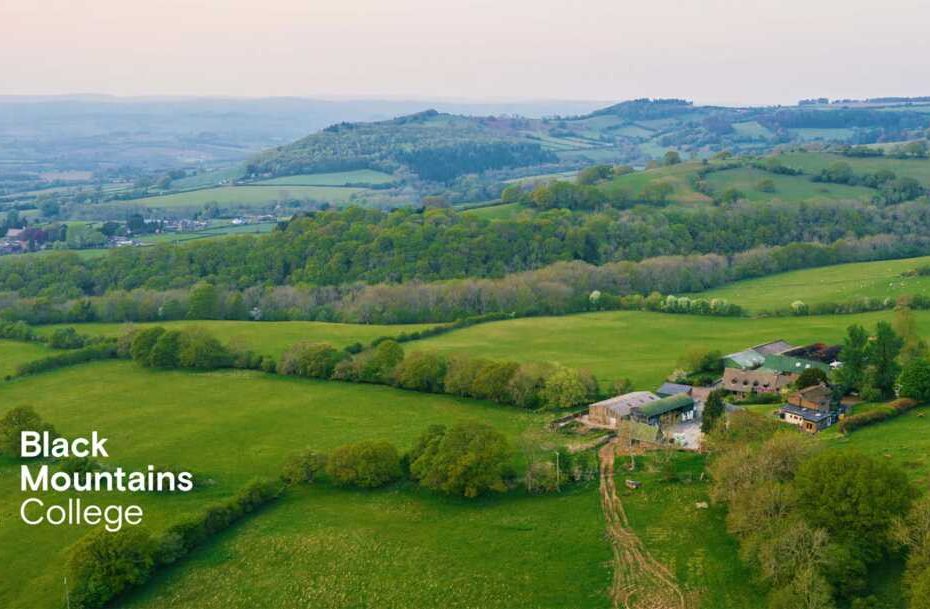 An aerial photograph of the Welsh countryside near Brecon and the Bannau Brycheiniog, with green fields and hedgerows, and a farm nestled in the foreground. The Black Mountains College logo is overlaid in white in the bottom left hand corner.