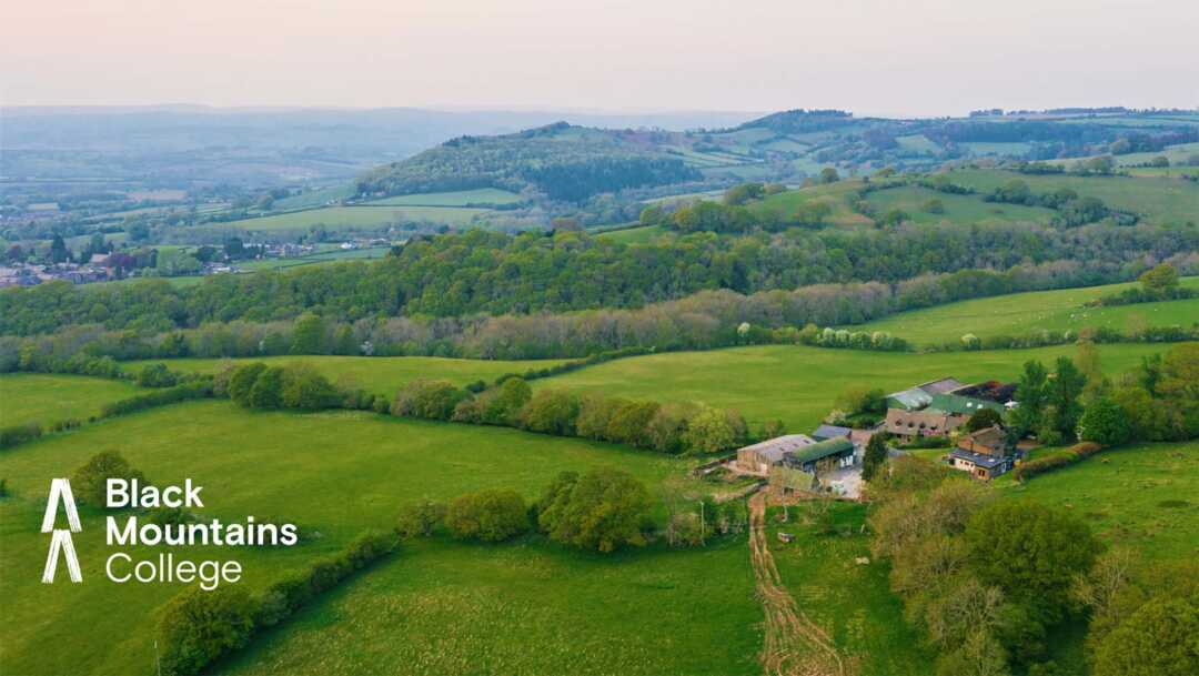 An aerial photograph of the Welsh countryside near Brecon and the Bannau Brycheiniog, with green fields and hedgerows, and a farm nestled in the foreground. The Black Mountains College logo is overlaid in white in the bottom left hand corner.