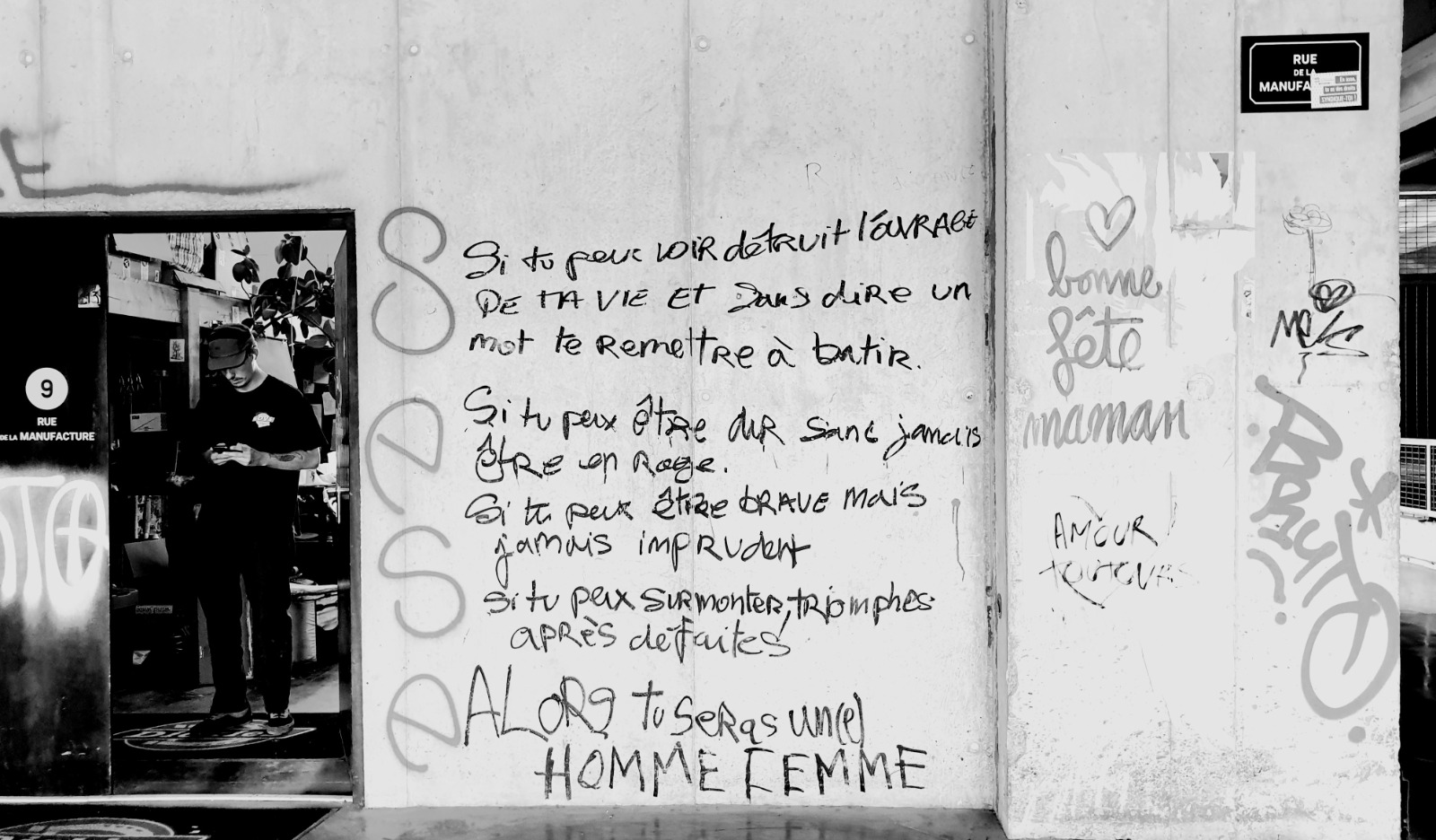 Black and white photo of a concrete wall filled with graffiti writing, and a man standing in an open doorway looking down at his phone.