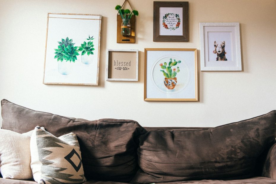 A saggy and comfortable looking brown sofa with cushions, under a collection of whimsical art and plants on the wall.