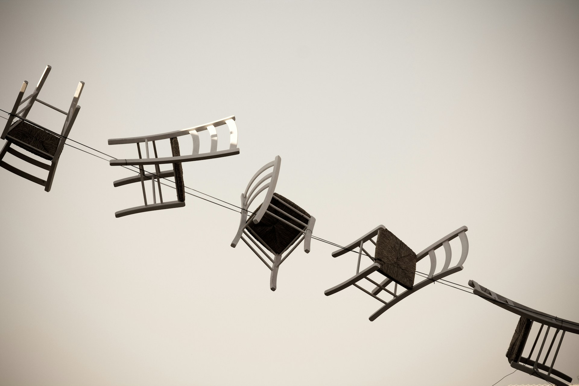 An arty black and white photo of 5 traditional wooden chairs with straw seats, strung along a wire like laundry, pointing in different directions.