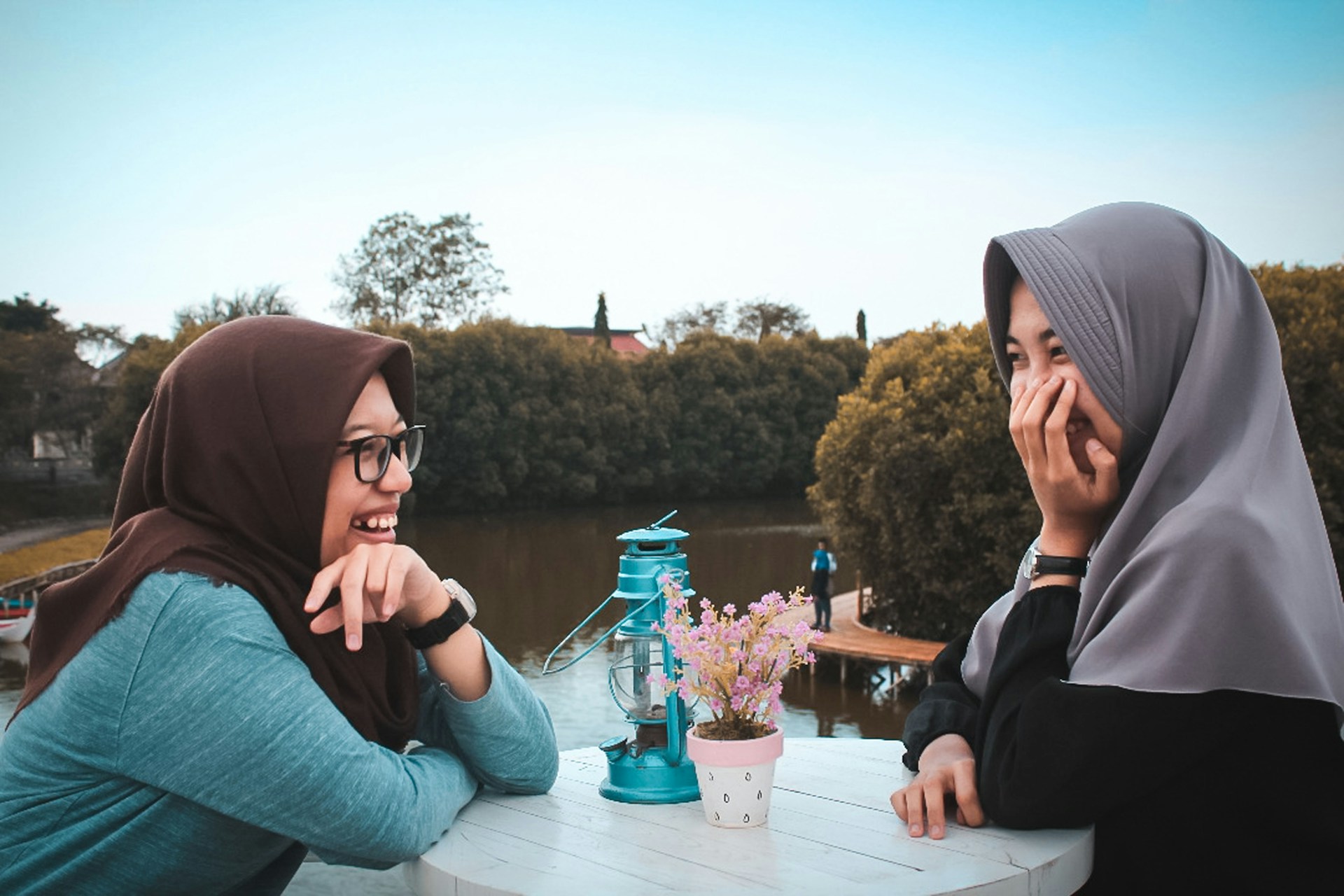 Two women are sat outdoors at a small table near a lake. They are talking and giggling. They both are wearing headscarves, long-sleeved tops and watches. One wears glasses. Between them on the table, a decorative lantern and a pot of flowers.