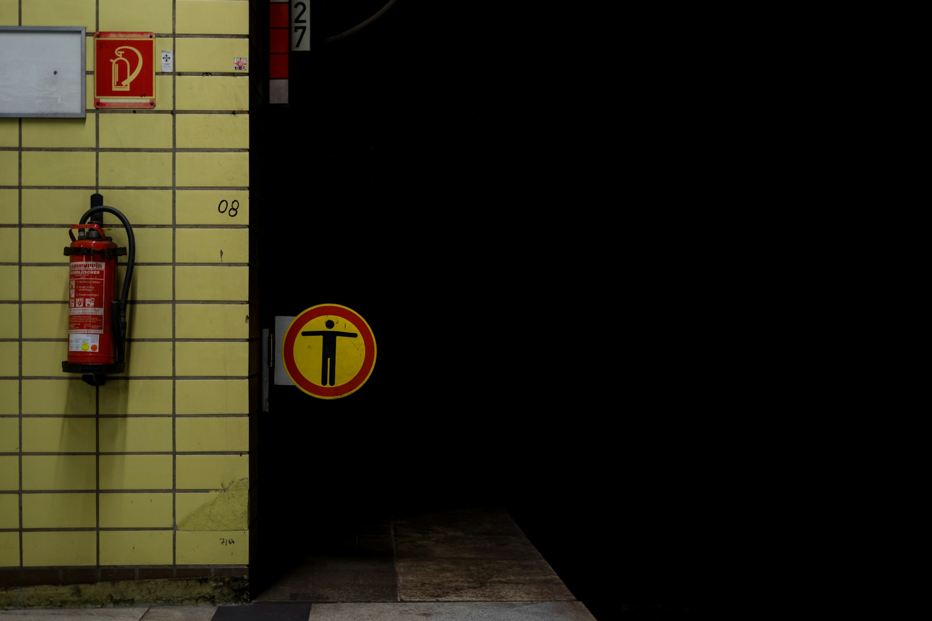 The entrance of a subway, underground, or metro tunnel. To the left of the shot, a wall tiled in very functional pale yellow rectangular tiles, with a fire extinguisher and signage. At the edge of the wall and above the end of the platform, a round alert sign sticks out, showing a person standing arms open inside a red circle on a yellow background. It means "no access". Behind the sign and to the right of the platform, the tunnel is nothing but darkness.