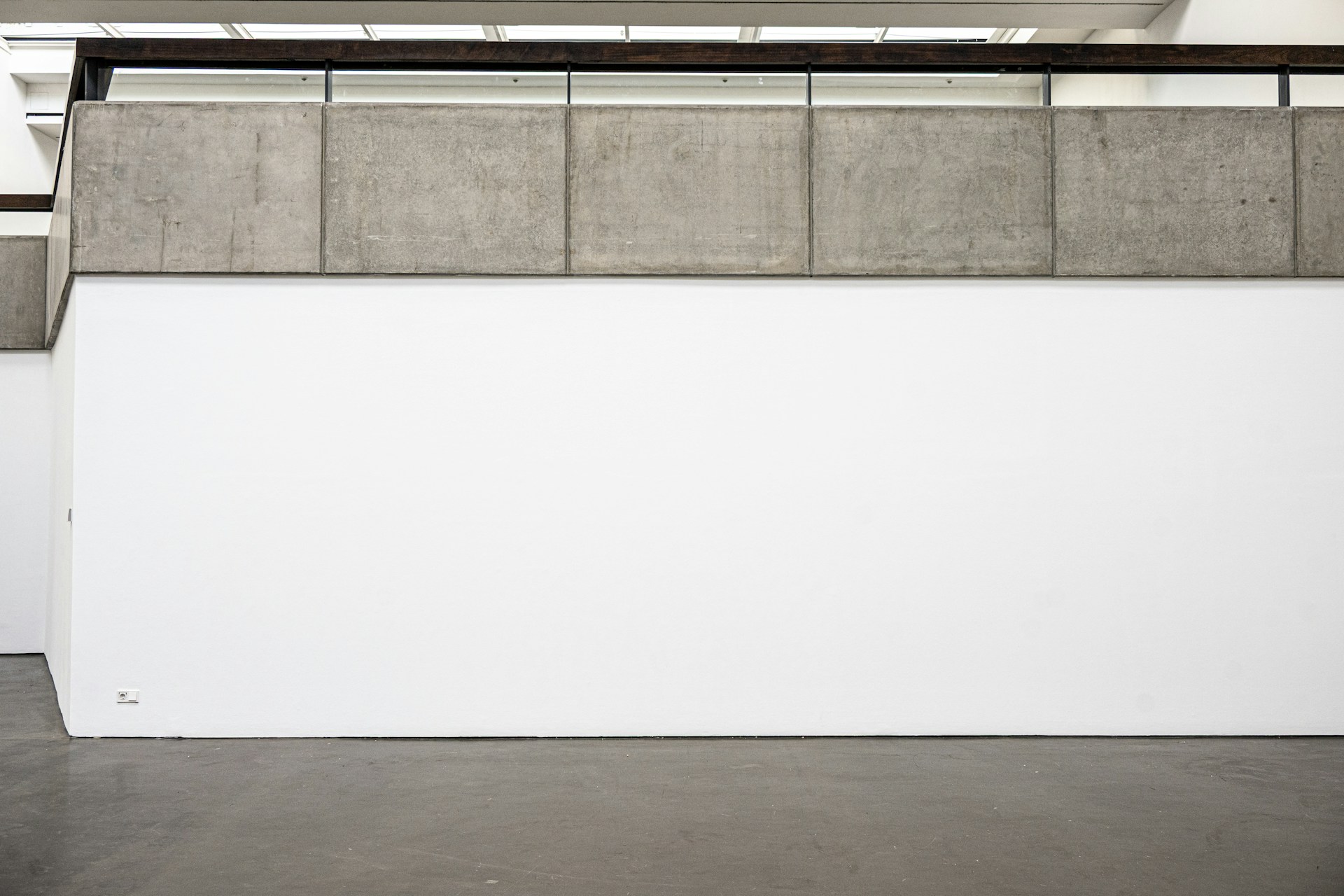 A white wall topped with concrete blocks and a black railing, under skylights.