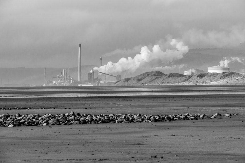 A black and white photograph of a wide, flat beach at low tide, with rocks in the foreground and the Port Talbot steelworks in the background, smokestacks billowing against a cloudy sky.