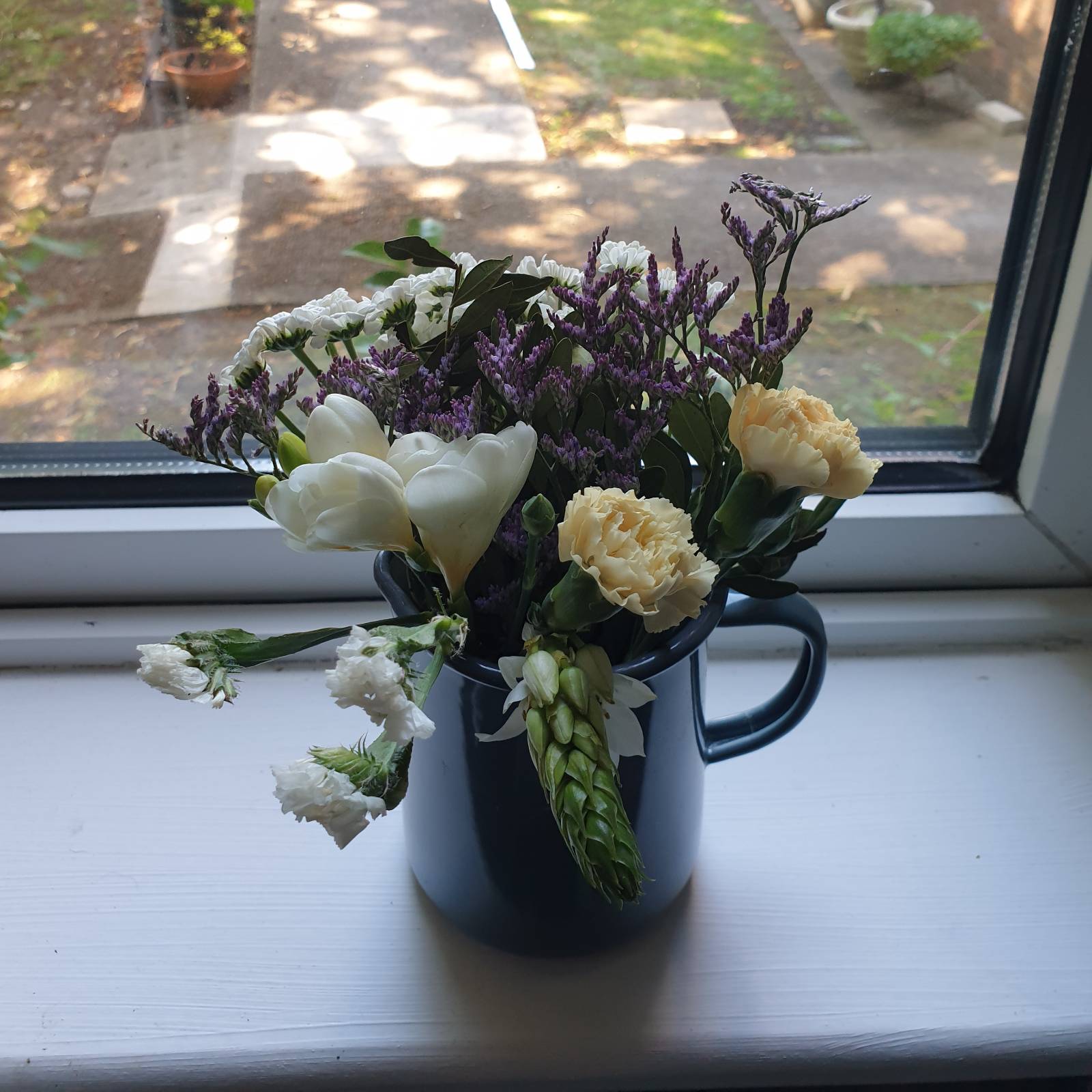 A small bouquet of white, purple and cream flowers sits in a grey enamel jug on a white window sill.