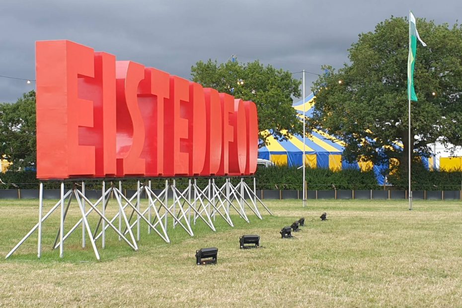 A shot of the big red "EISTEDDFOD" letters on the Eisteddfod field. At their feet, spotlights to light them up when it's dark. In the background, trees and a yellow and blue striped tent.