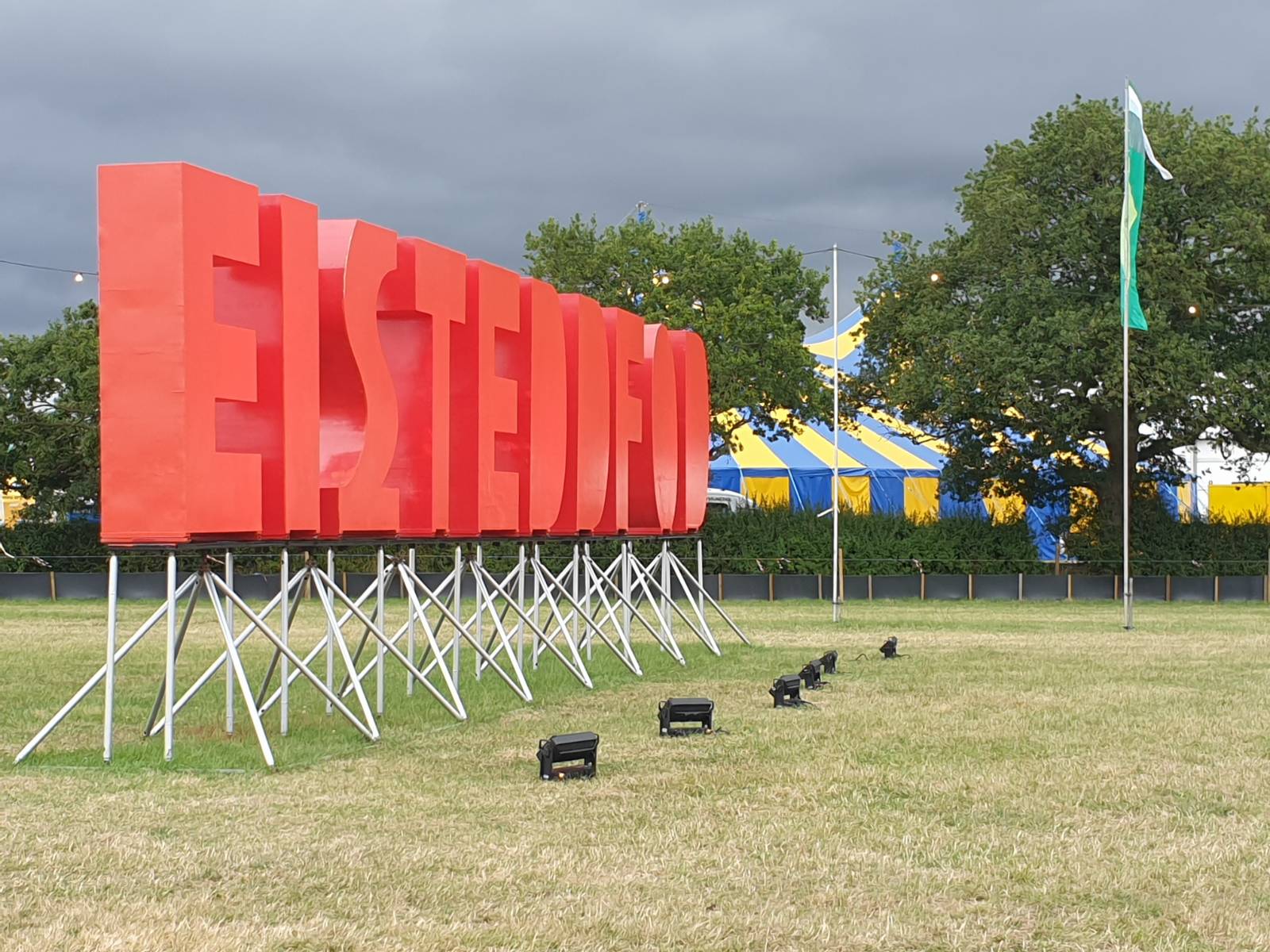 A shot of the big red "EISTEDDFOD" letters on the Eisteddfod field. At their feet, spotlights to light them up when it's dark. In the background, trees and a yellow and blue striped tent.