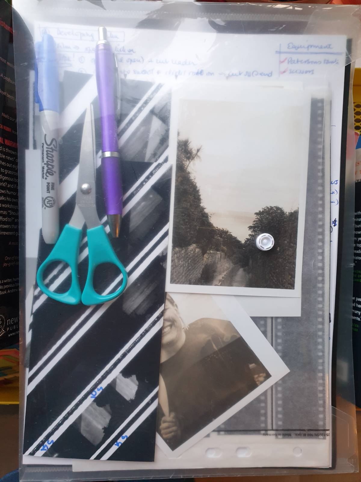 A transparent plastic folder contains a ballpen, a marker and a pair of scissors, as well as film negatives, some contact sheet test strips, and some prints of a landscape and you can see part of a portrait too.