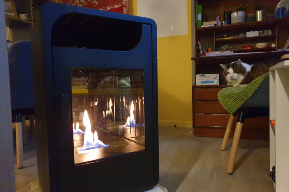A black standalone stove is lit up with yellow flame inside. Behind it, the cat sits on his chair, and there is a bureau with papers, pens and plants.
