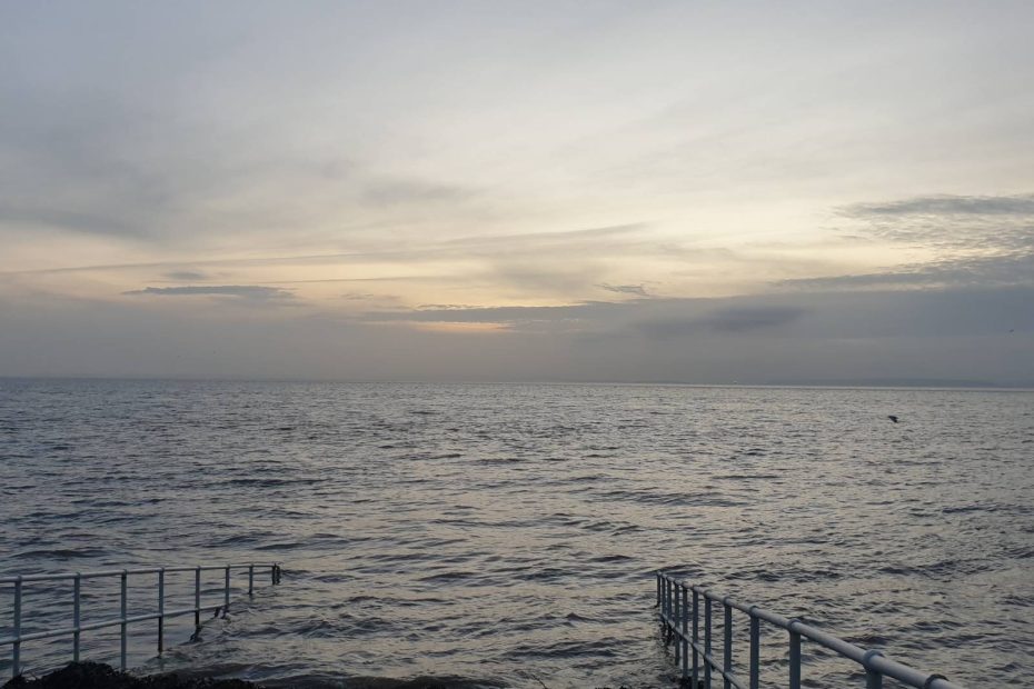 The sea at Penarth at high tide at dawn, looking south from the RNLI lifeboat station. There's cloud cover on the horizon and the dawn light above that.