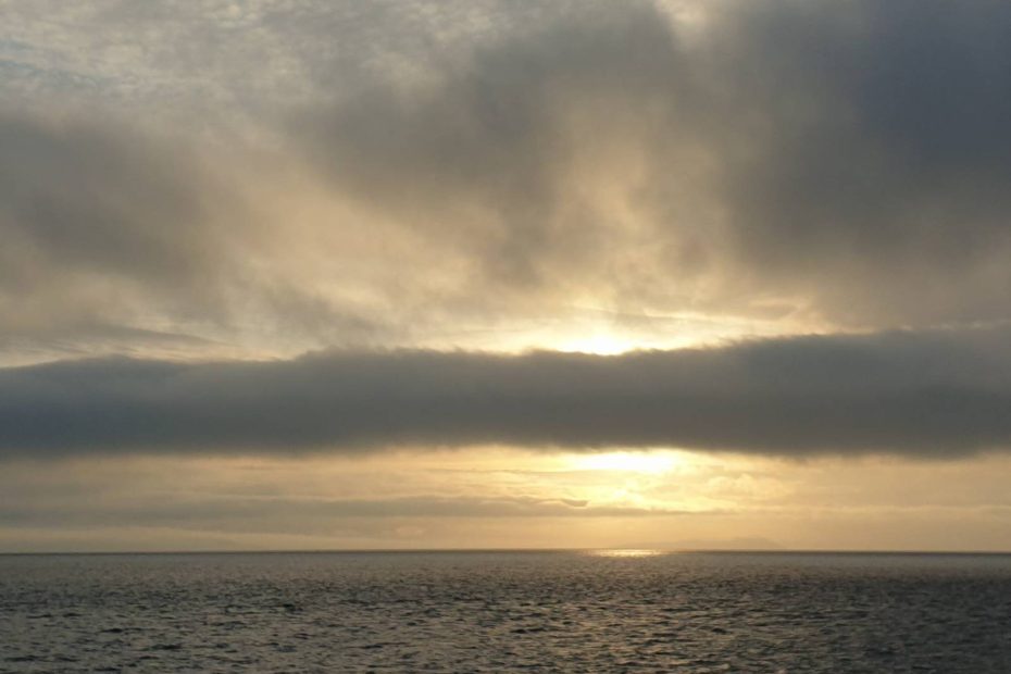 The sea with a band of cloud in the sky, behind which a golden sunrise is glowing.