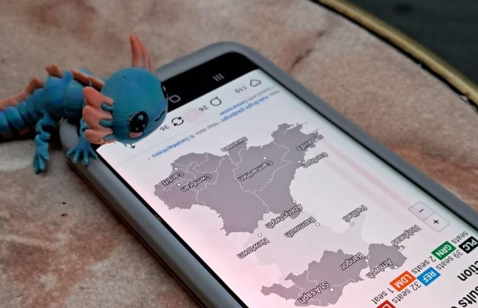 A small blue articulated plastic axolotl, perched on the edge of a phone - on the screen is a map of Wales with partial Senedd election results.