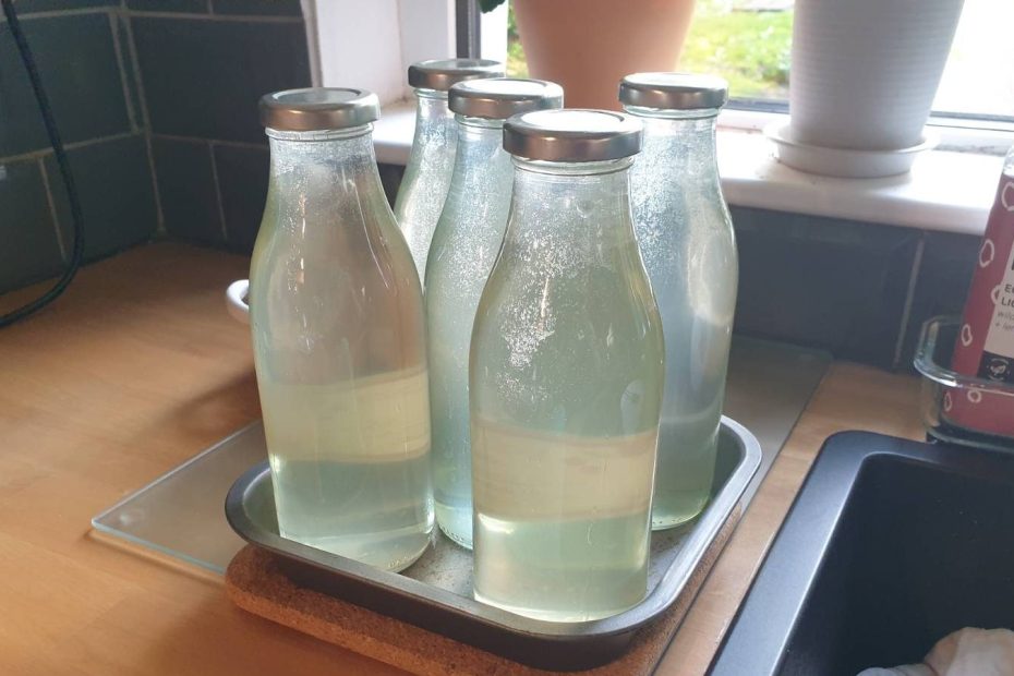 Four glass pint bottles filled with freshly made elderflower cordial, cooling on the side of the sink.