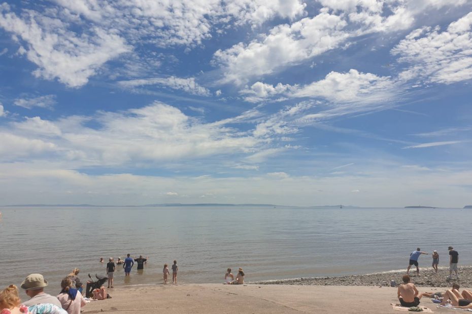 The slipway at Penarth beach, there are people in the water and sitting in the sun, and the sky is dotted with interesting clouds. The sea is very tranquil.