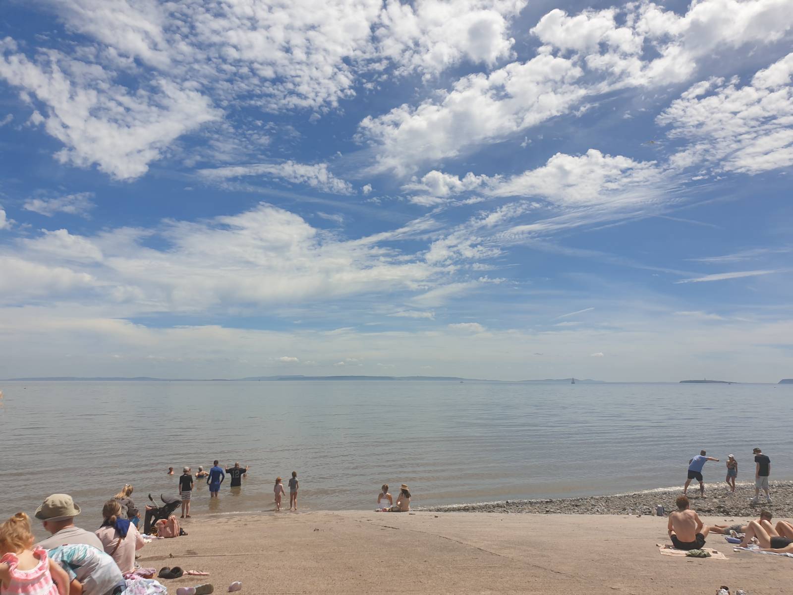 The slipway at Penarth beach, there are people in the water and sitting in the sun, and the sky is dotted with interesting clouds. The sea is very tranquil.