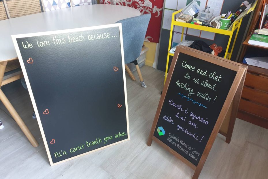 A flat blackboard reads in English and in Welsh "we love this beach because..." with space for people to write their thoughts in chalk, and an A-board blackboard reads in English and in Welsh "come and chat to us about bathing water!" with the Natural Resources Wales logo hand drawn at the bottom, it's all a bit wonky and home made haha.