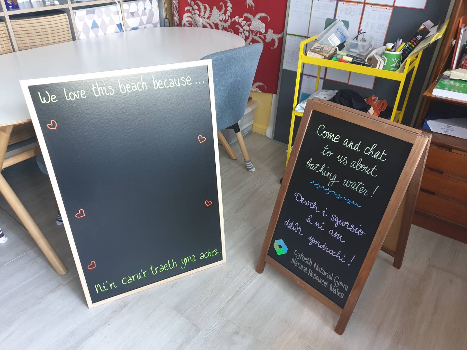A flat blackboard reads in English and in Welsh "we love this beach because..." with space for people to write their thoughts in chalk, and an A-board blackboard reads in English and in Welsh "come and chat to us about bathing water!" with the Natural Resources Wales logo hand drawn at the bottom, it's all a bit wonky and home made haha.