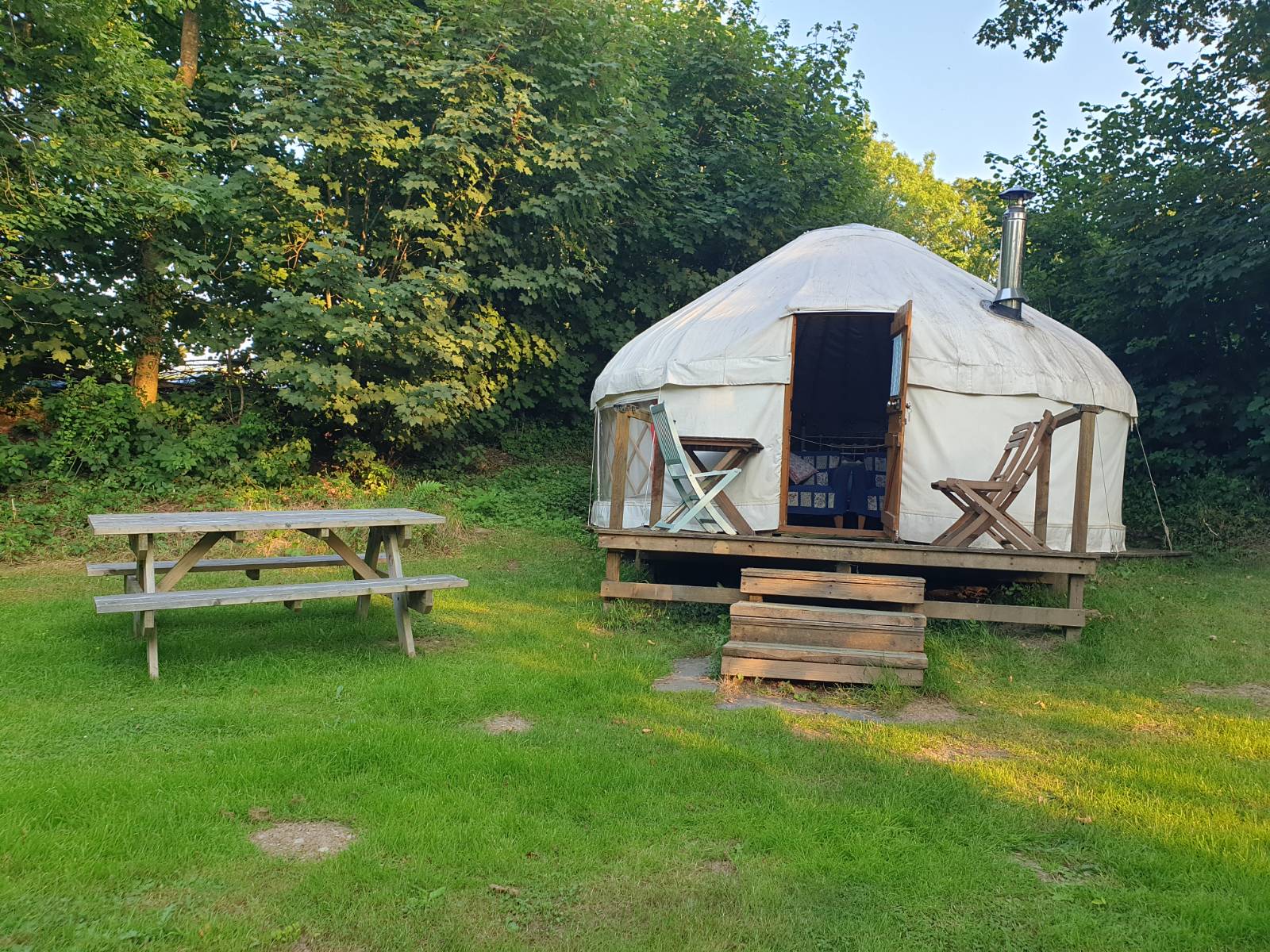 A yurt in a glade, surrounded by trees with chairs on the patio and a picnic bench on the grass in front.