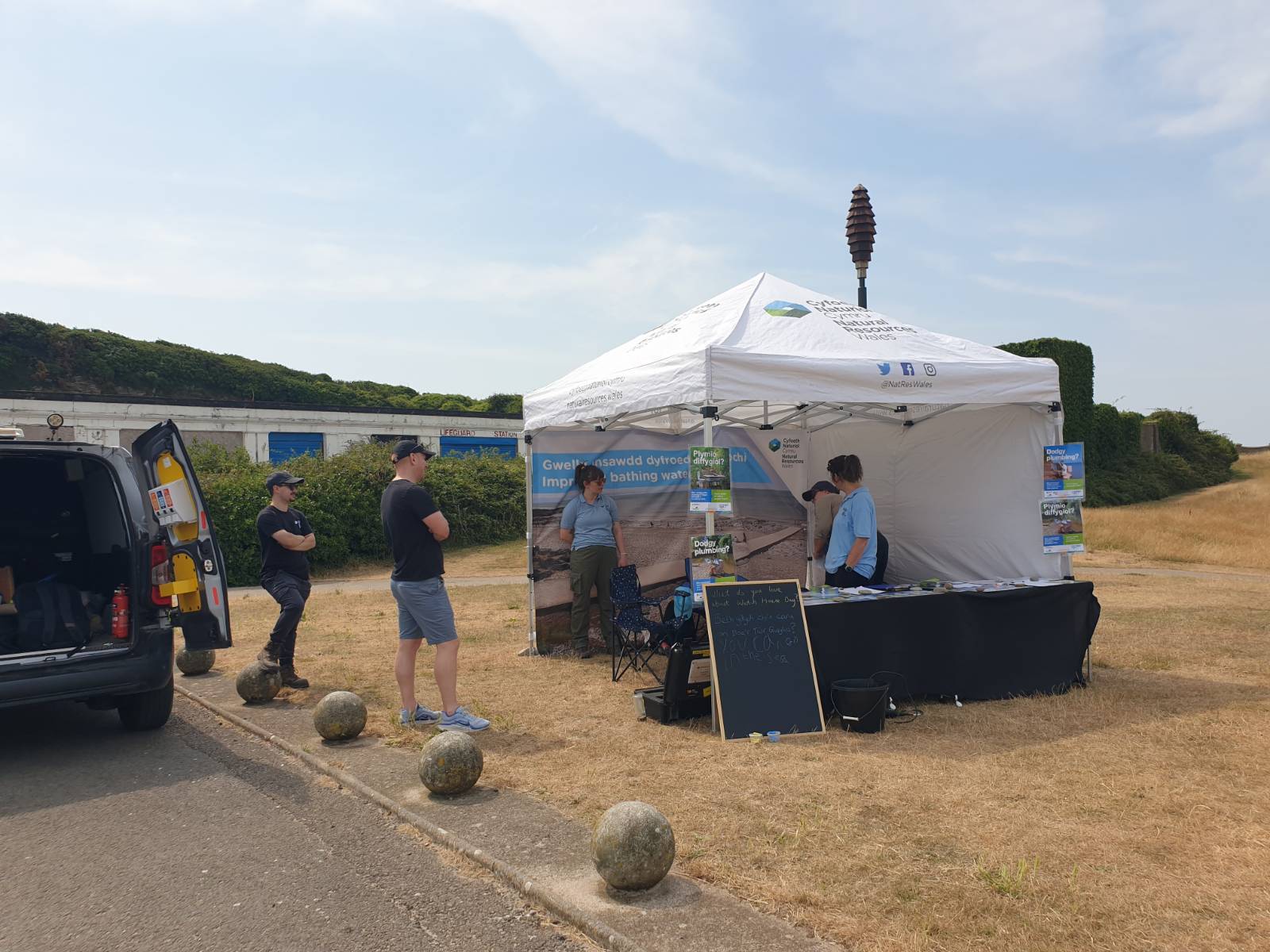 A pop-up stand on an expanse of very dry yellow grass. It's a gazebo that says "Natural Resources Wales", and it has a long table with papers and displays, and bits of kit to do with measuring water quality. There are also posters and an illustration of the beach on the back panel of the gazebo. Various members of staff are standing around and in the gazebo, and nearby there is a work van with its doors open.
