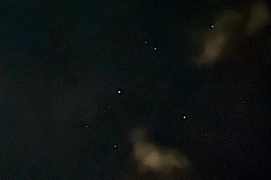 A shot of a night sky with blurry clouds, the pinpricks of many stars, and a little line which is a satellite moving across the sky during the length of the camera exposure.