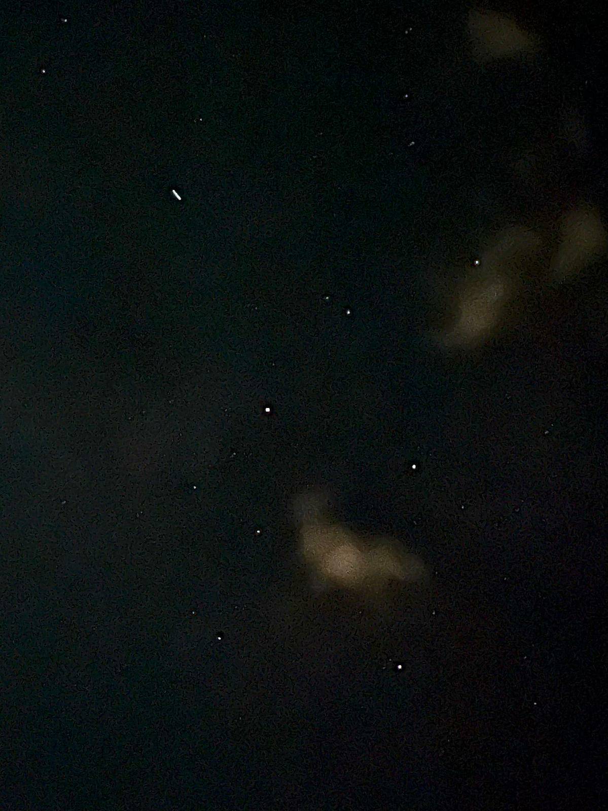 A shot of a night sky with blurry clouds, the pinpricks of many stars, and a little line which is a satellite moving across the sky during the length of the camera exposure.