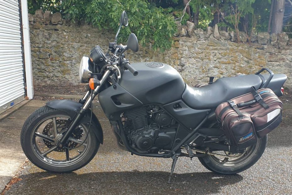 A black motorbike with panniers parked in front of a wall and some garages. .