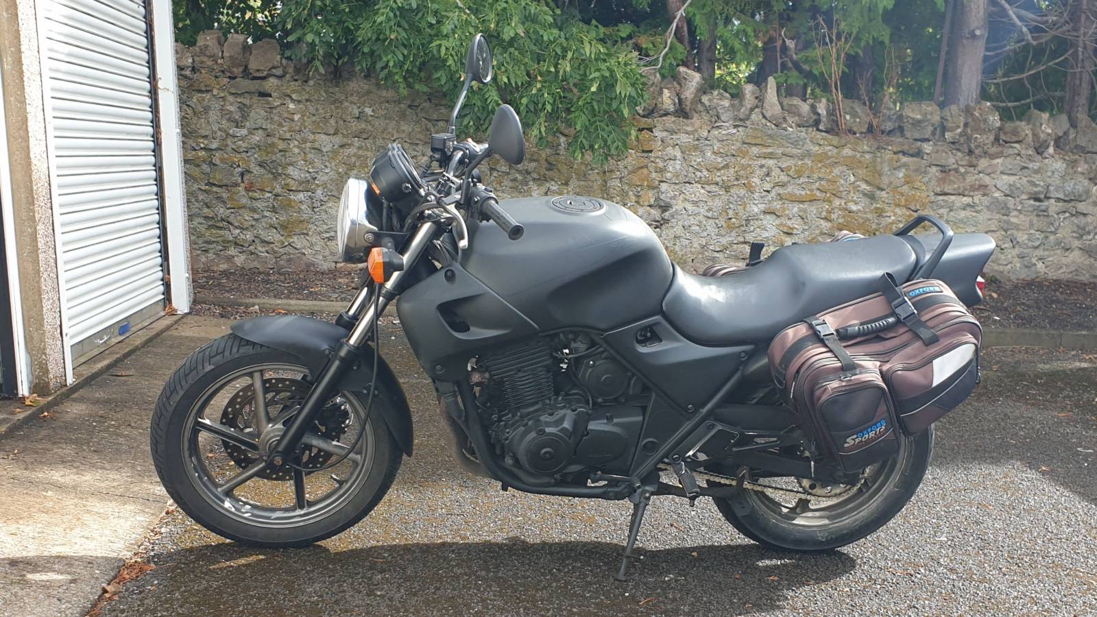 A black motorbike with panniers parked in front of a wall and some garages.