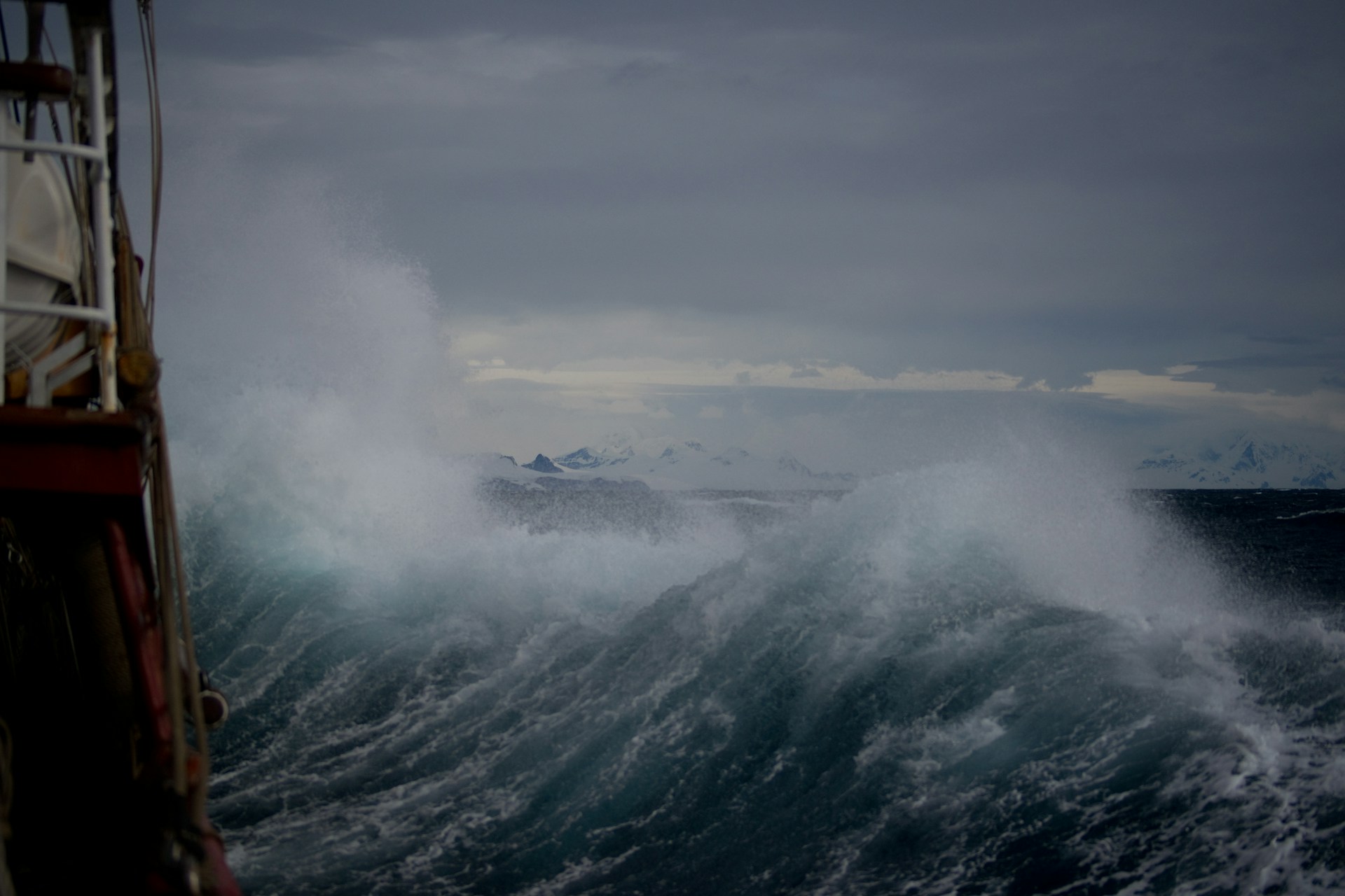 A stormy sea full of big waves and spray, seen from the deck of a ship in the midst of it. There is land in the distance.