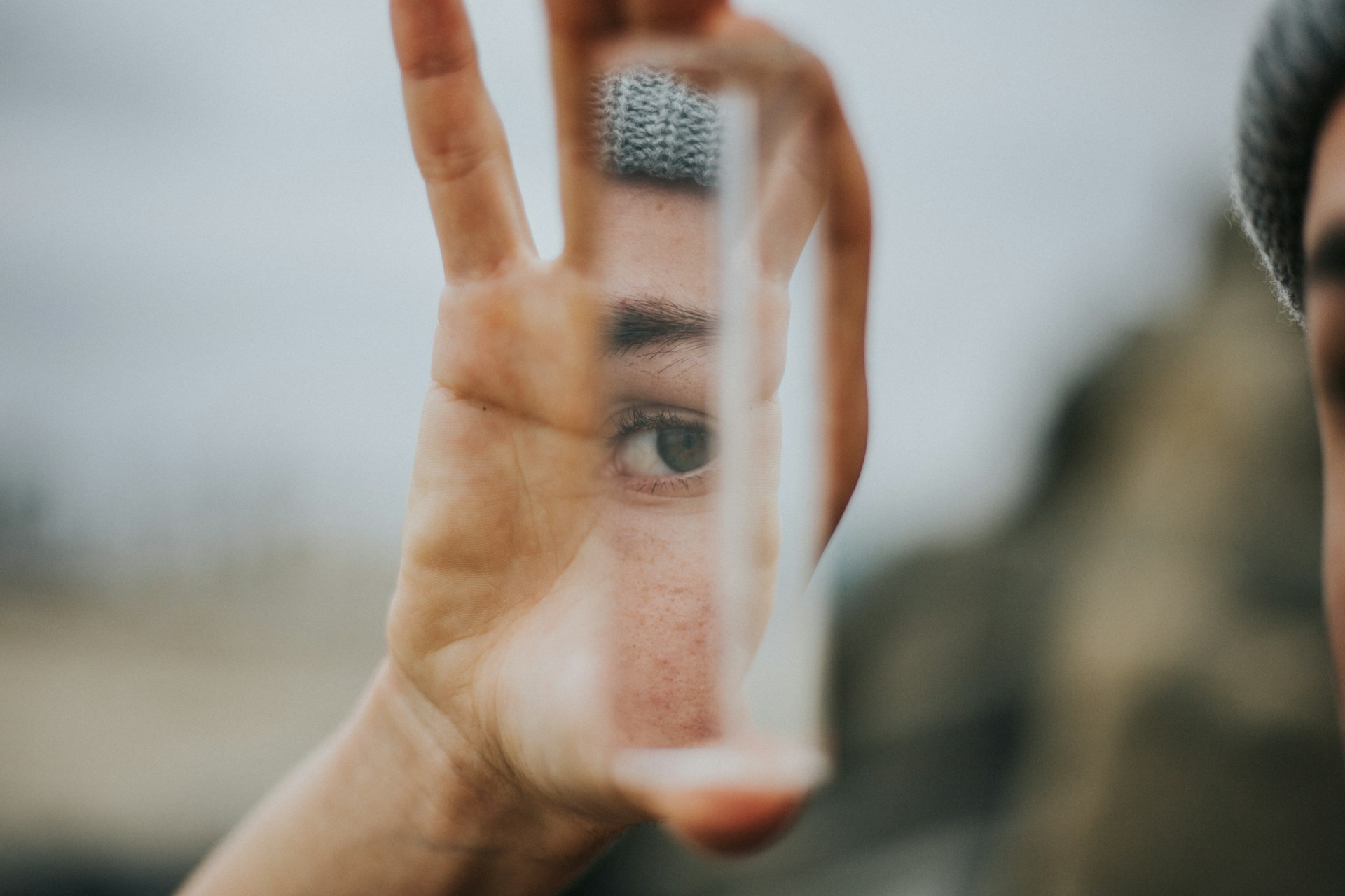 A close-up photograph of a person holding a shard of glass in front of their face. Their eye is visible and in sharp focus through the glass, while the rest of the image is blurred. The effect suggests looking through a lens or prism, with the glass both clarifying and distorting the view.