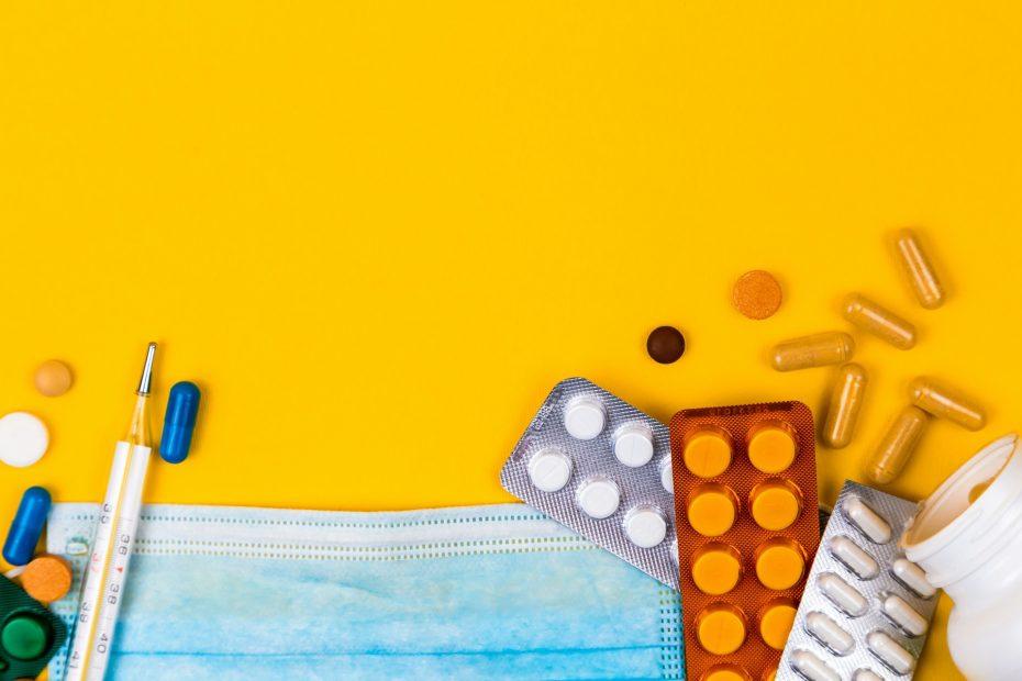 A photo of medicines in blister packs and loose pills and capsules, a mercury thermometer and a clinical mask, against a deep yellow background.