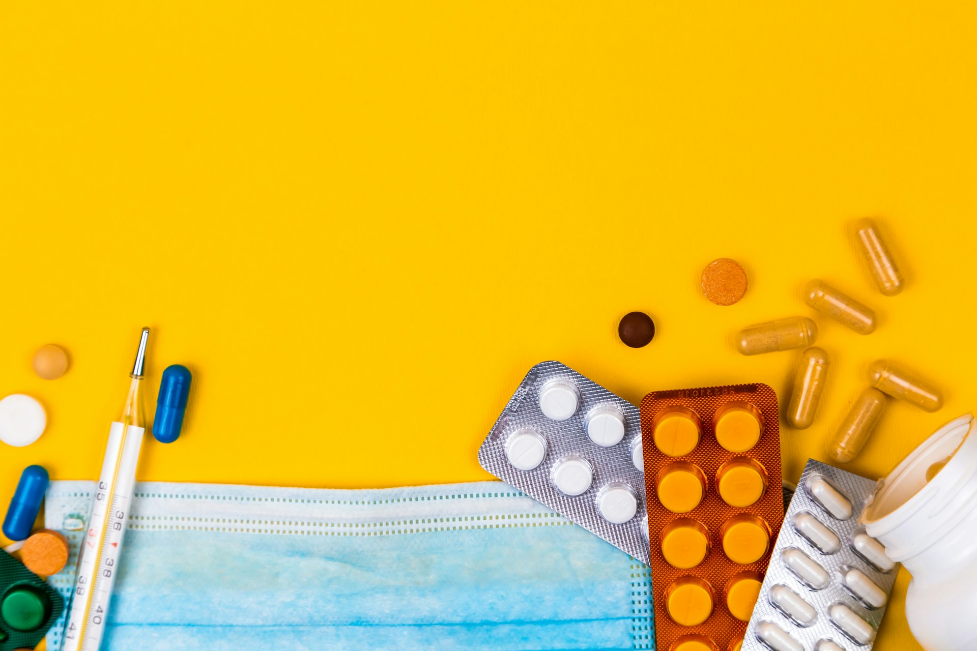 A photo of medicines in blister packs and loose pills and capsules, a mercury thermometer and a clinical mask, against a deep yellow background.